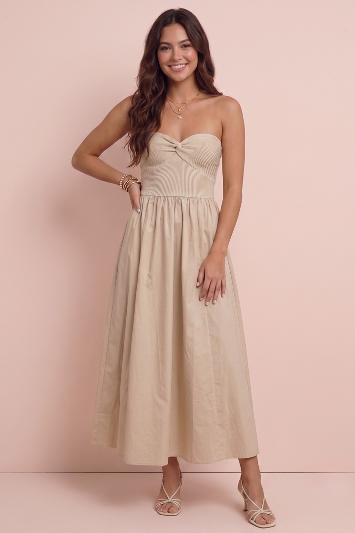 A woman modeling a strapless midi dress in a light beige or cream color, standing against a solid pale pink studio backdrop.