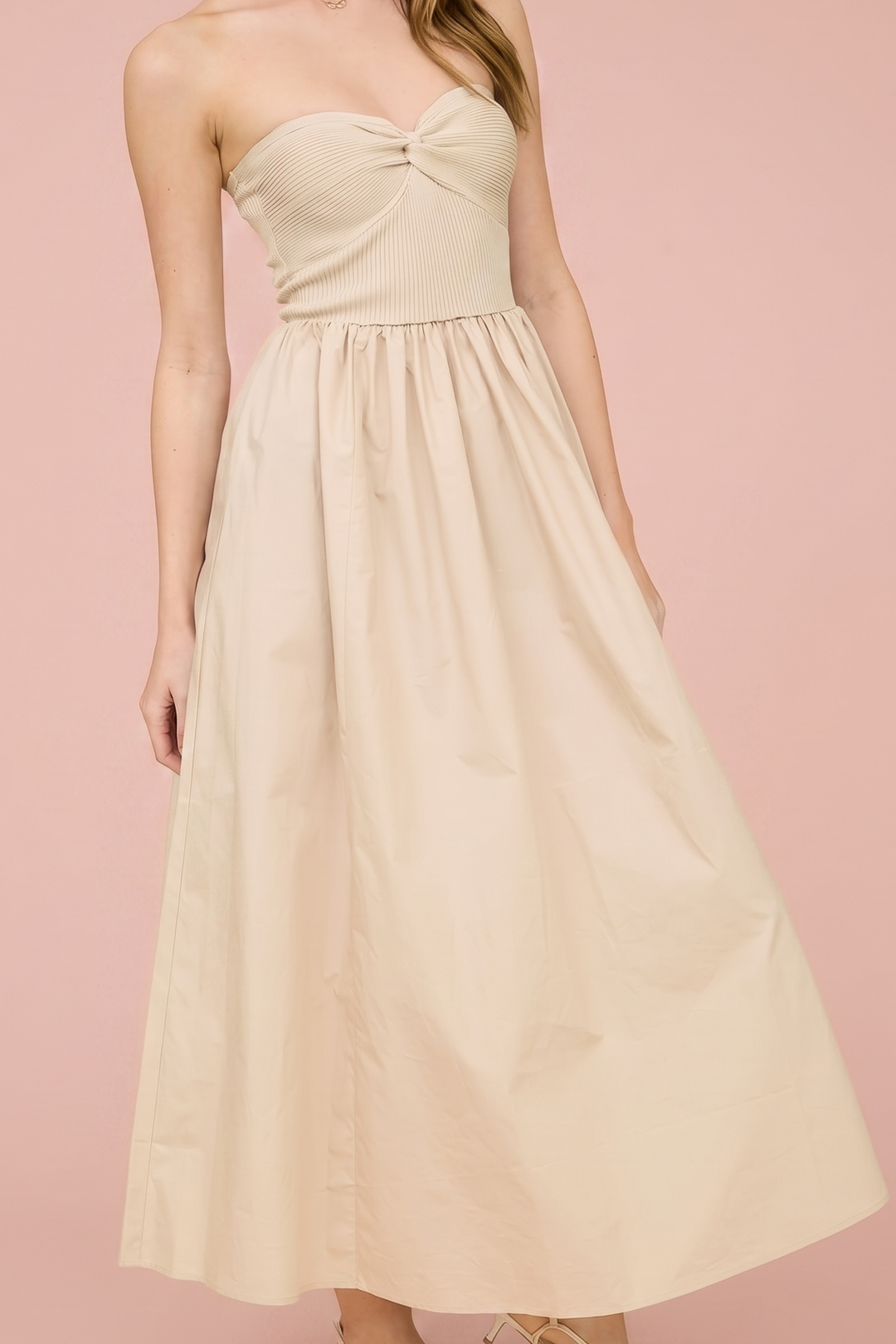 A woman modeling a strapless midi dress in a warm cream or beige color, standing against a solid dusty pink studio background.