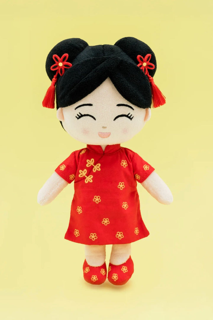 A plush, cultural doll with a black wig, red floral dress, and red shoes, smiling cheerfully against a yellow background.