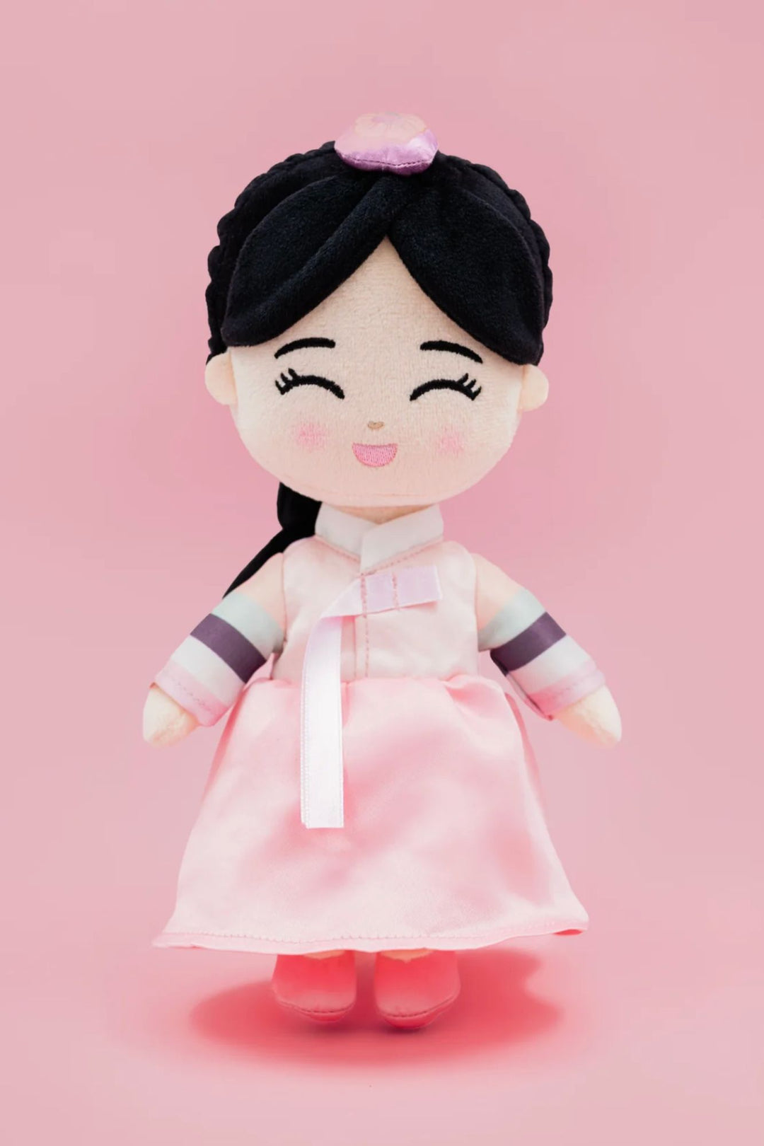 A plush doll with a smiling face, wearing a pink hanbok dress and a black headband with a floral accent, against a pink background.
