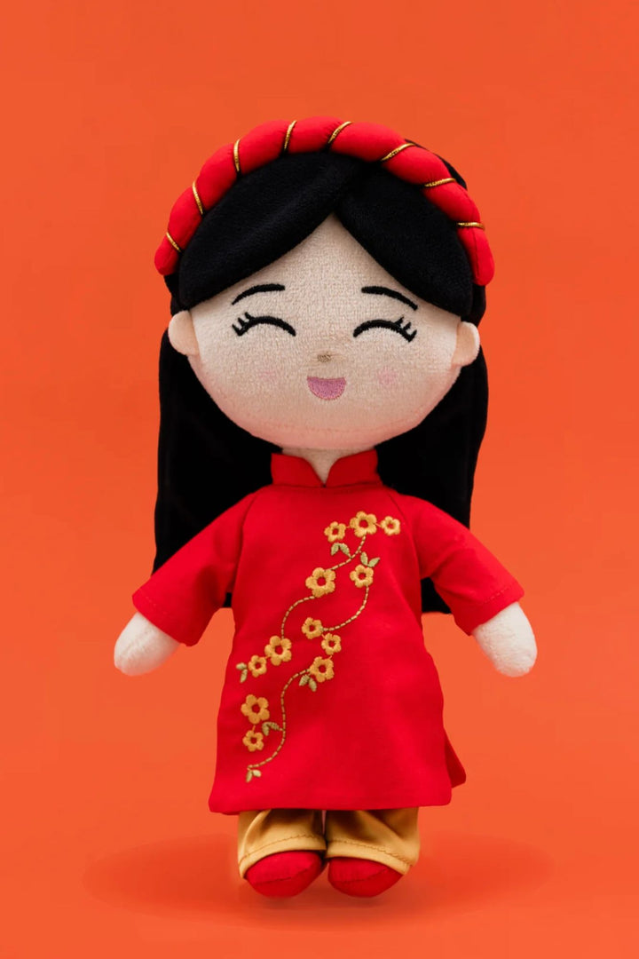 Plush doll with dark hair, red dress with gold floral embroidery, and red shoes, against vibrant orange background.