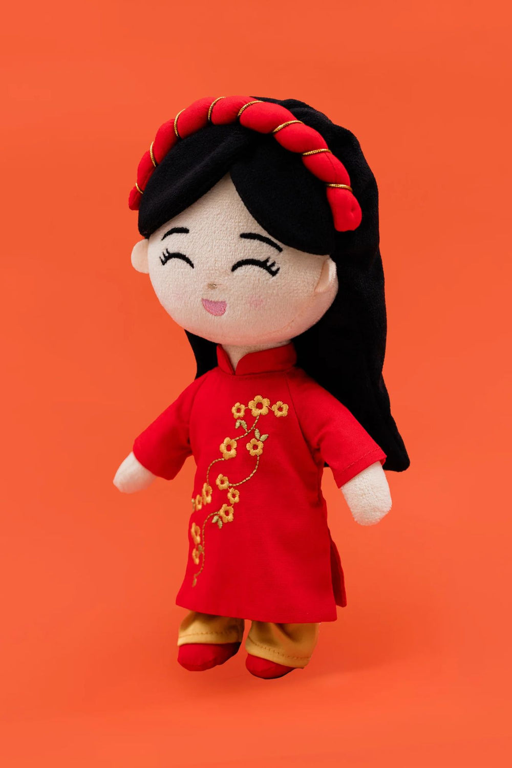 A smiling plush doll wearing a red dress with gold flower designs, black hair styled in a bun, and a cheerful expression on its face.