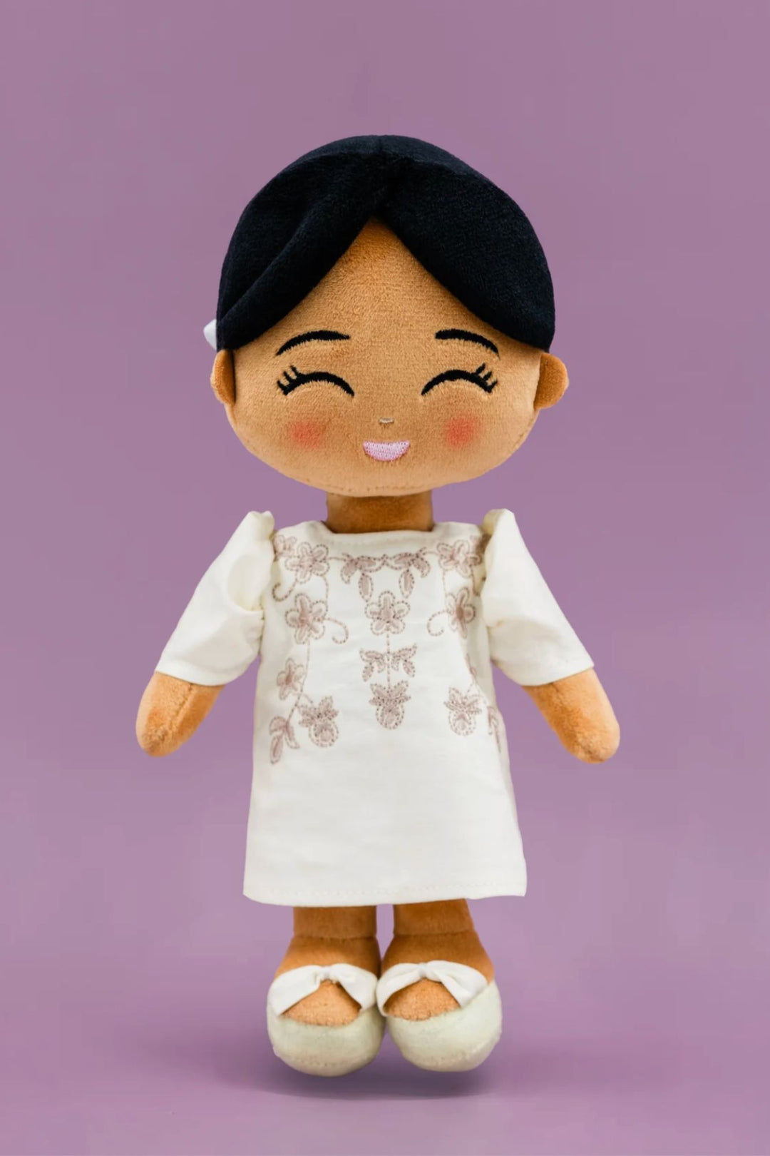 A plush, smiling doll with black hair, wearing a white dress with floral embroidery, and white and orange shoes.
