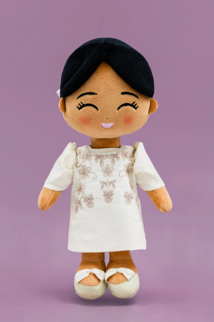 A plush, smiling doll with black hair, wearing a white dress with floral embroidery, and white and orange shoes.