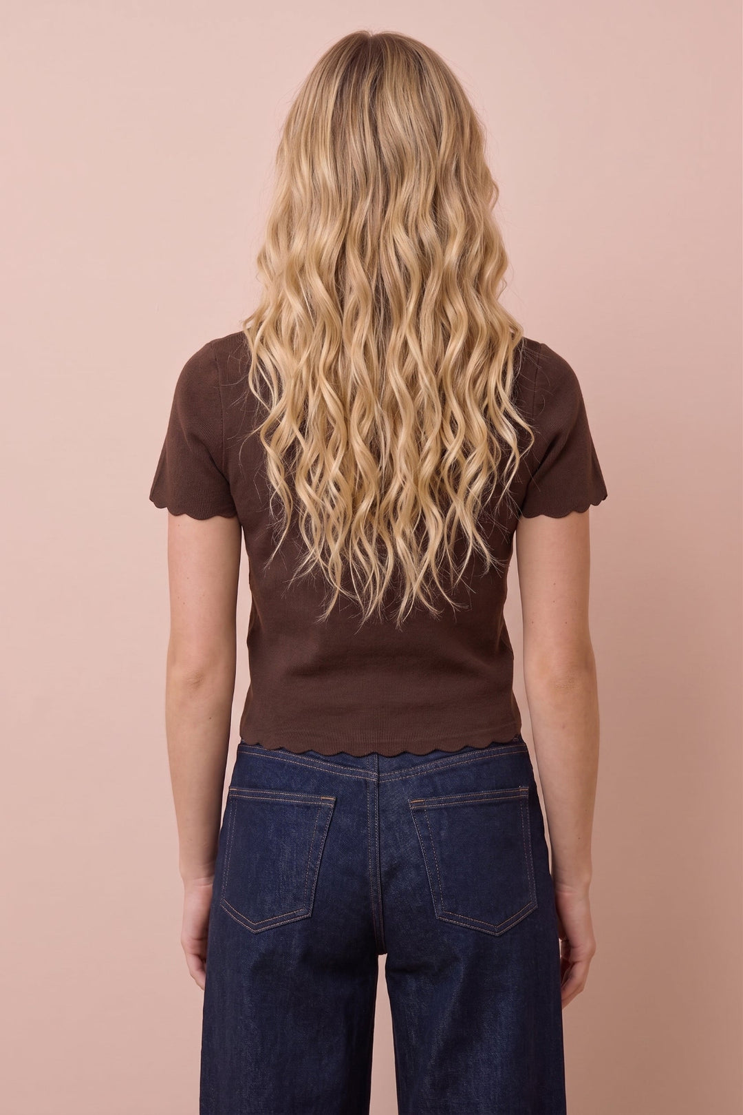 A woman viewed from behind wearing a dark brown short-sleeve top with scalloped edges and dark blue jeans, standing against a solid pale pink background.