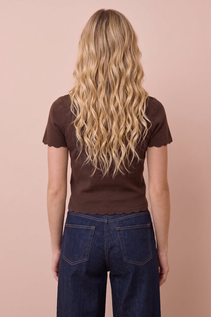 A woman viewed from behind wearing a dark brown short-sleeve top with scalloped edges and dark blue jeans, standing against a solid pale pink background.
