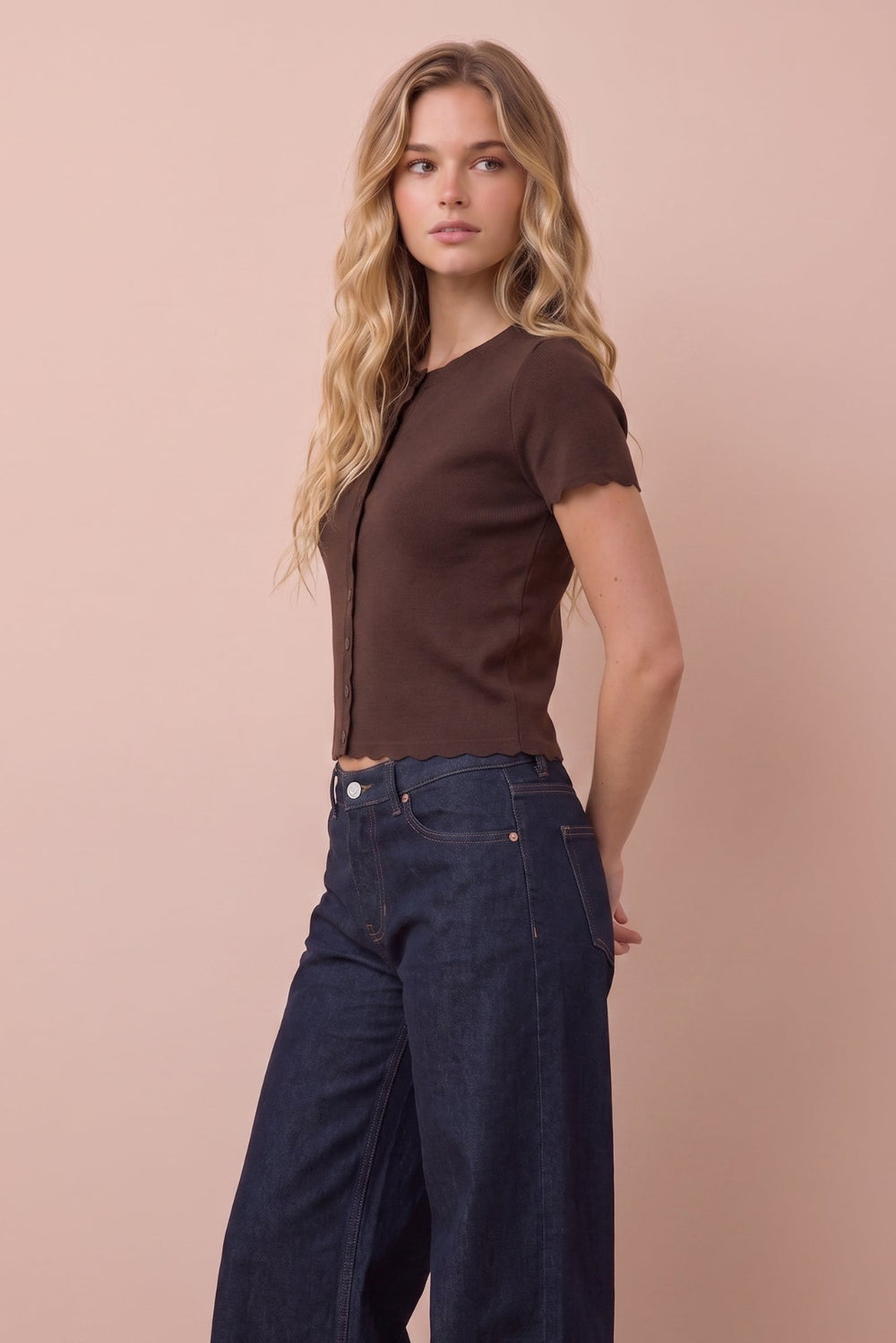A woman modeling a short-sleeve brown button-front top and dark wide-leg jeans against a solid pale pink background.