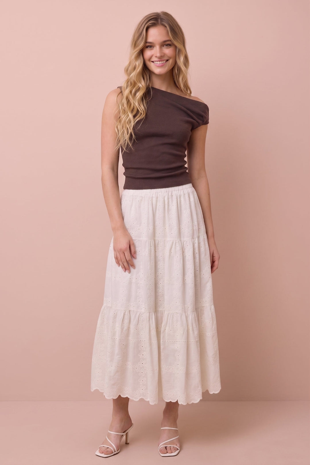 A woman modeling a dark brown off-the-shoulder knit top and a cream-colored, tiered tiered maxi skirt against a solid light pink studio background.