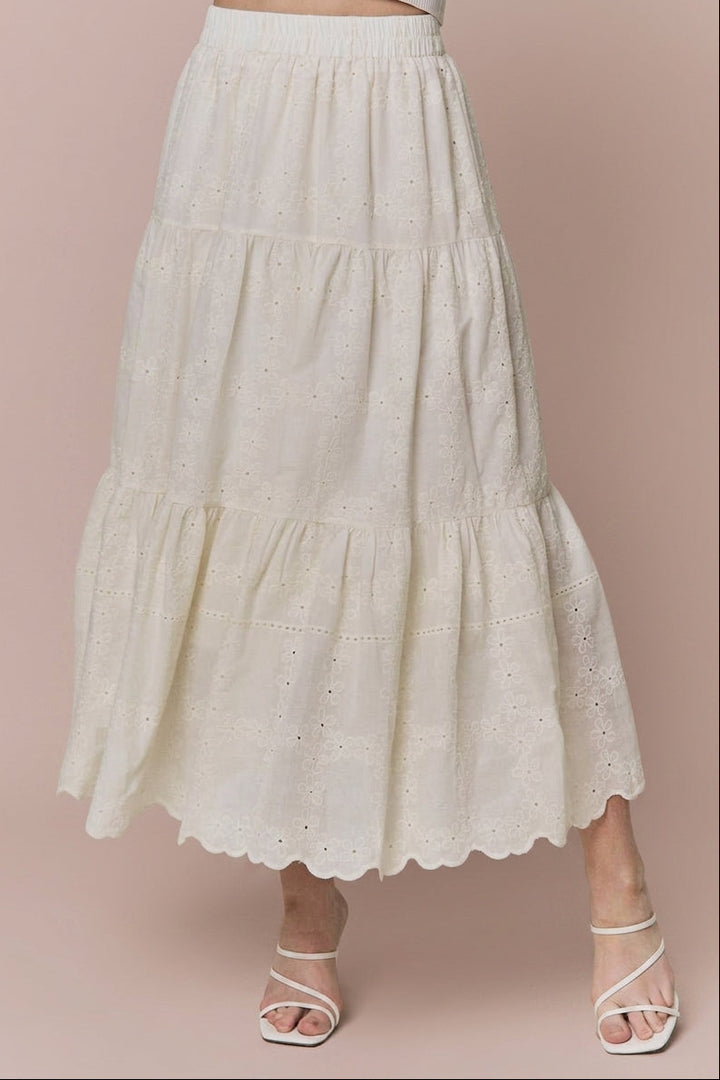 An off-white tiered eyelet skirt with a scalloped hem, shown on a model wearing white strappy heels against a solid dusty pink background.