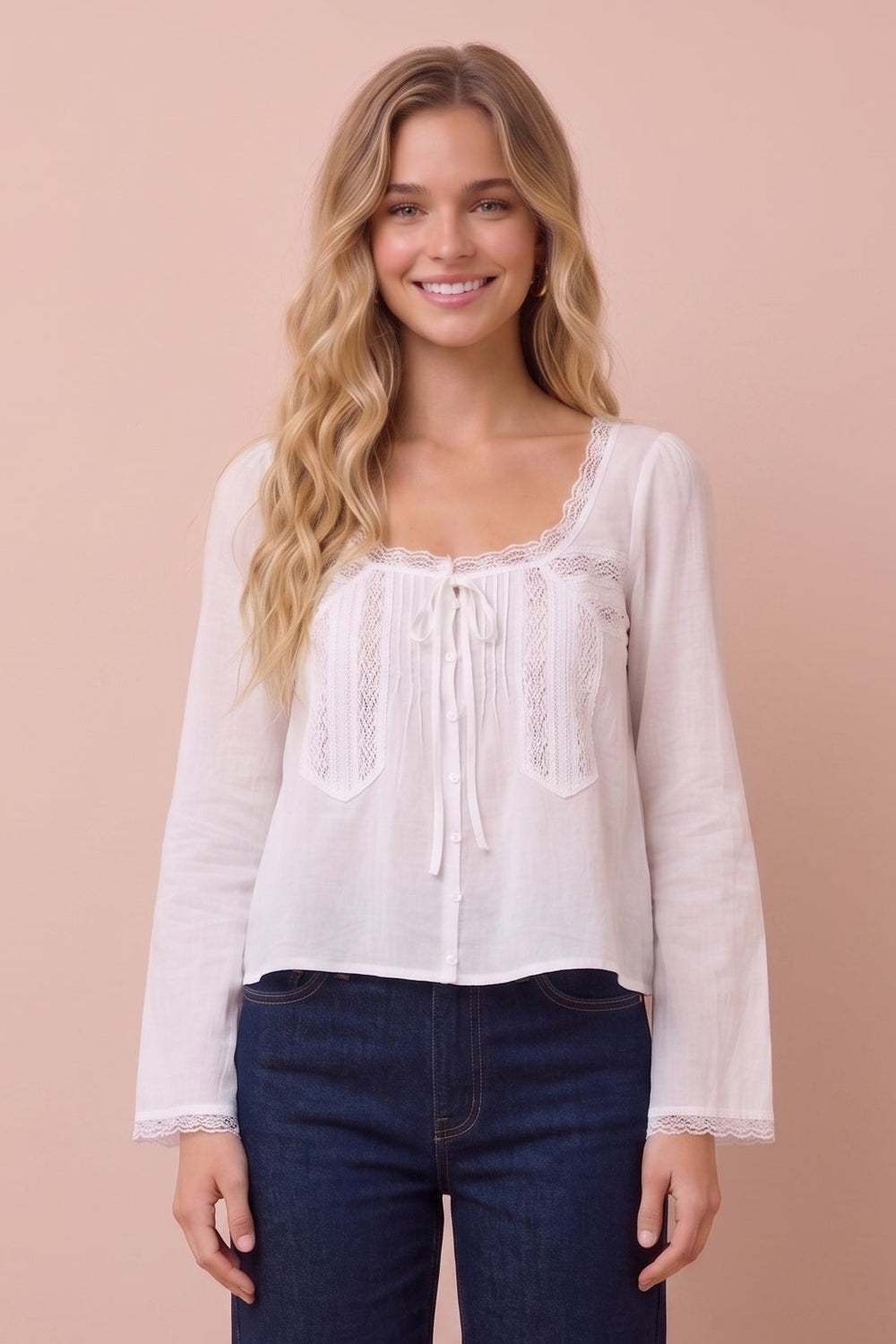 A woman stands against a solid pale peach studio background wearing a white long-sleeve lace-trim blouse and dark blue wide-leg jeans.