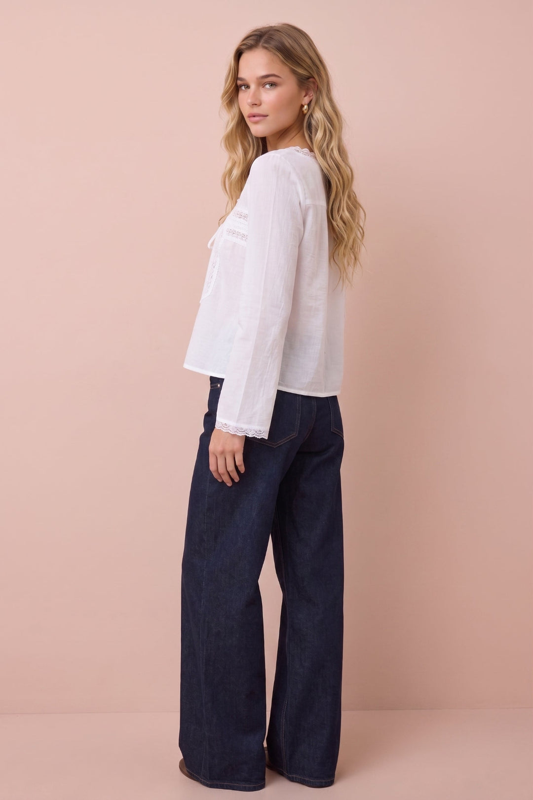 A woman modeling a white long-sleeve blouse with lace trim and dark wide-leg jeans, standing against a solid pale pink studio background.
