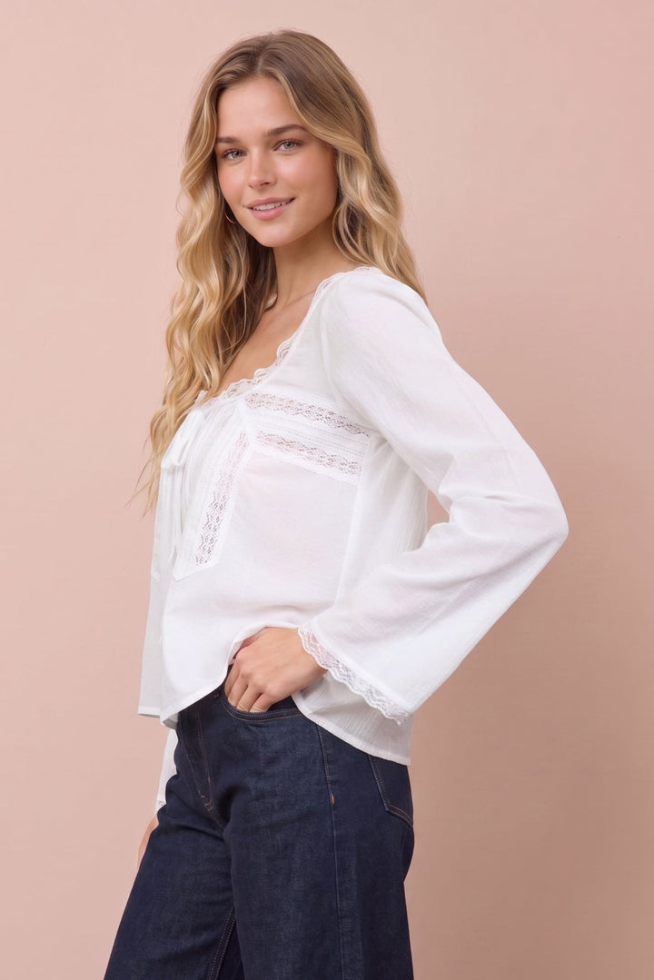 A woman modeling a white long-sleeve blouse with lace trim and dark wide-leg jeans against a solid pale pink studio background.