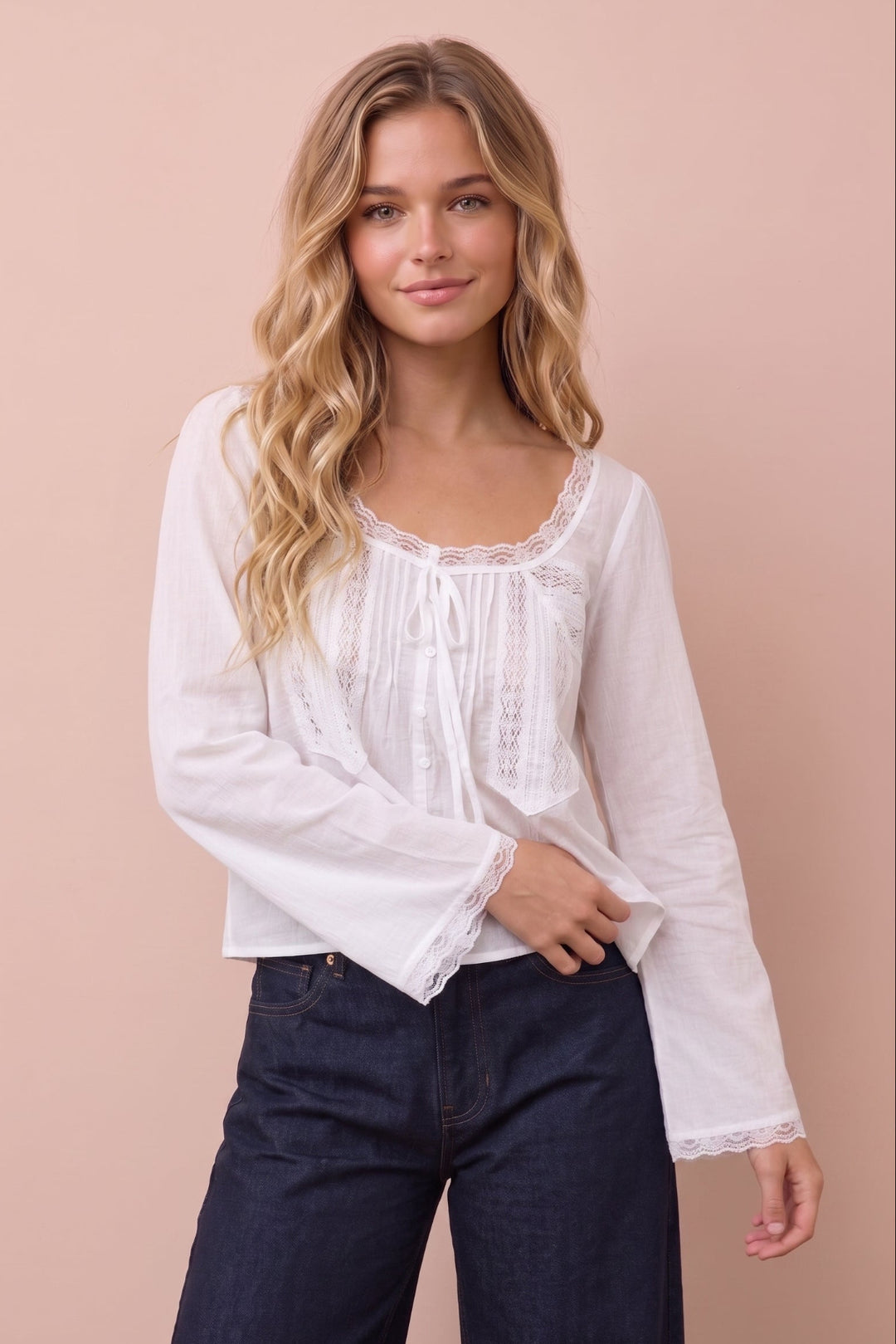 A woman modeling a white long-sleeve blouse with lace trim and a tie neckline, paired with dark blue jeans, against a solid pale pink background.