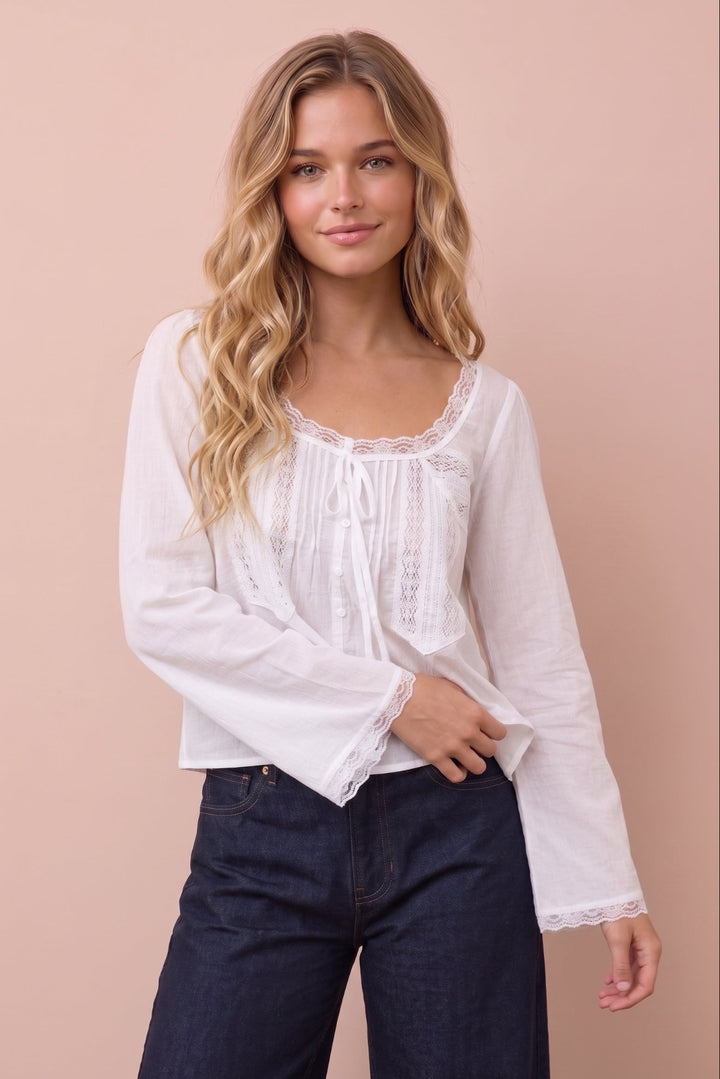A woman modeling a white long-sleeve blouse with lace trim and a tie neckline, paired with dark blue jeans, against a solid pale pink background.