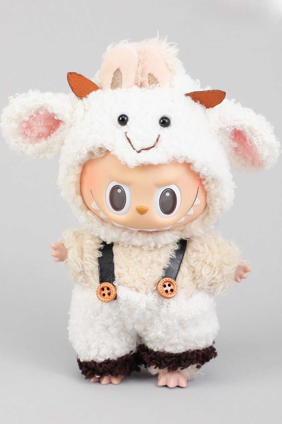 Plush white sheep toy with large expressive eyes, orange ears, and suspender-style straps on fluffy brown-and-white outfit.