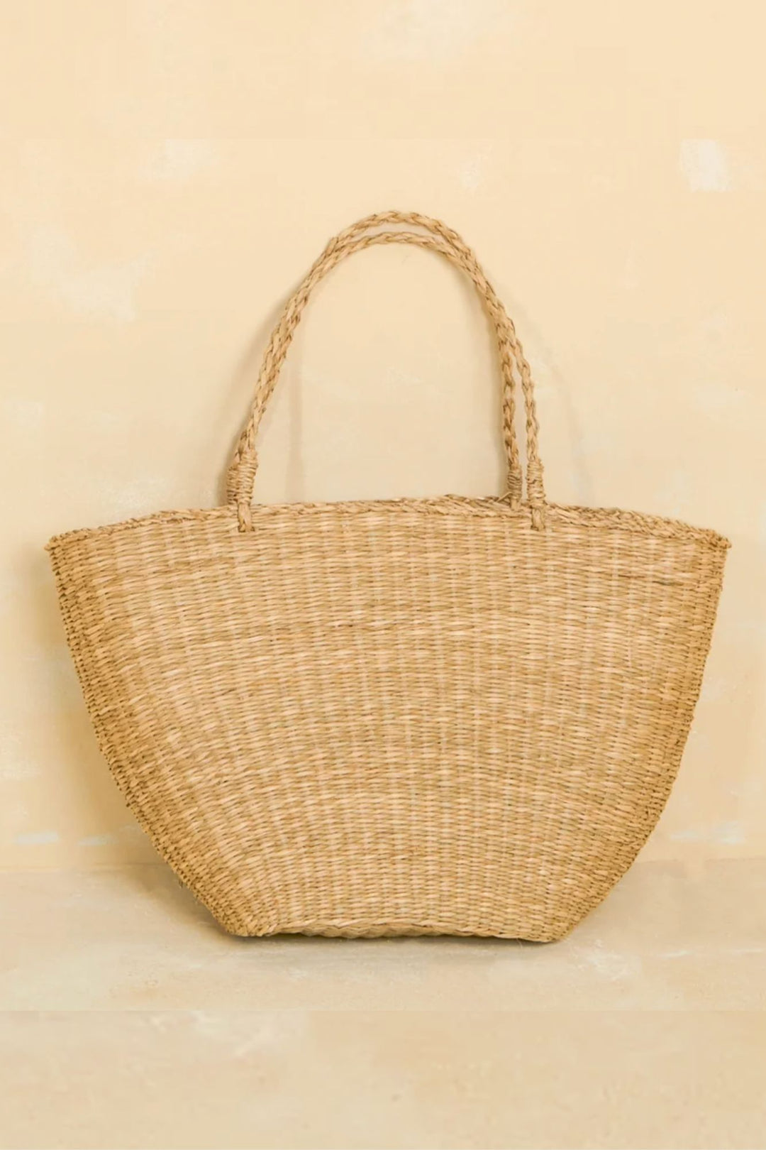 A beige, handwoven straw tote bag with a sturdy, tight-knit design and plaited handles, showcasing natural, eco-friendly craftsmanship.