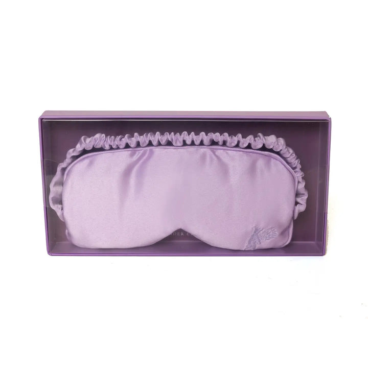 A lavender-colored satin eye mask with a detachable pouch filled with cassia seeds and dried lavender flowers, designed for a calming and relaxing sleep experience.