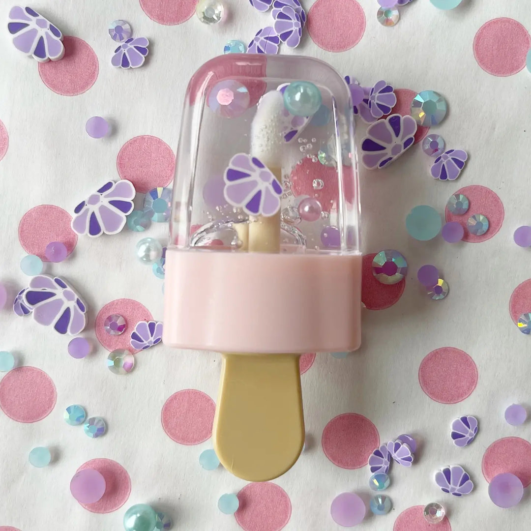 A pink and purple popsicle-shaped lip gloss applicator surrounded by colorful floral and polka dot-shaped cosmetic embellishments.