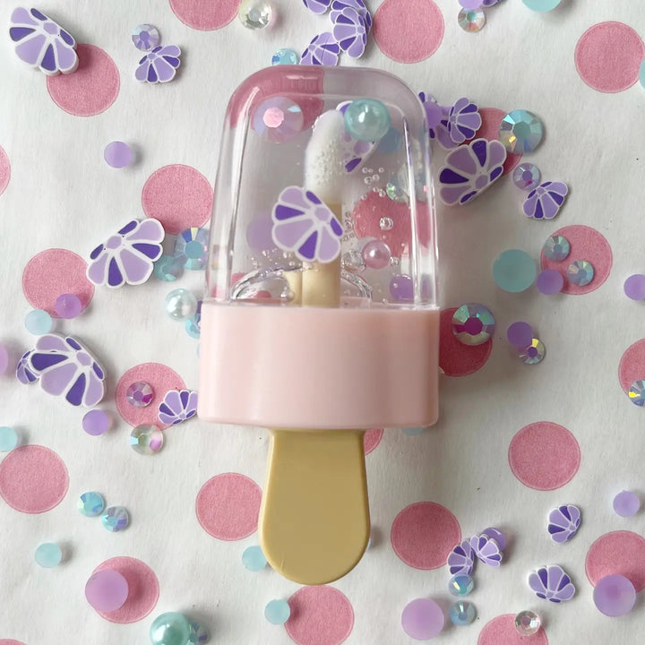 A pink and purple popsicle-shaped lip gloss applicator surrounded by colorful floral and polka dot-shaped cosmetic embellishments.