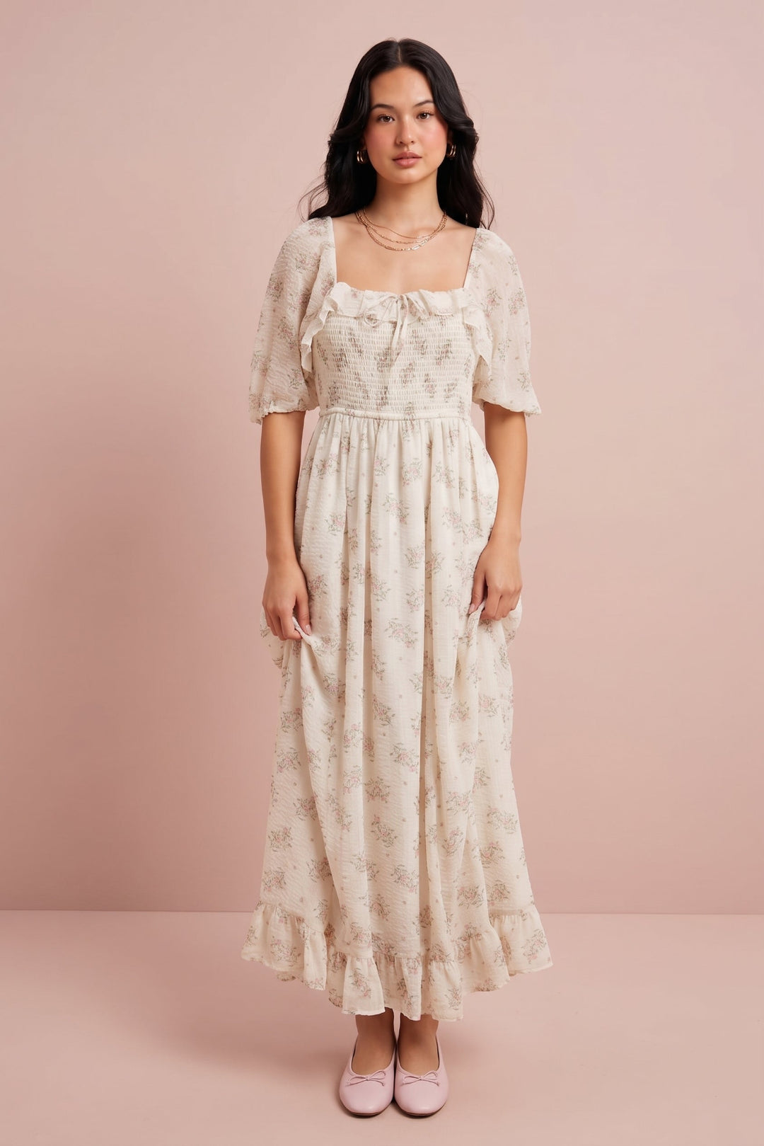 A woman stands against a soft pink studio backdrop wearing a cream maxi dress with a subtle floral print, short puff sleeves, a square neckline, and a gathered waist. She is also wearing pale pink flats and layered necklaces.