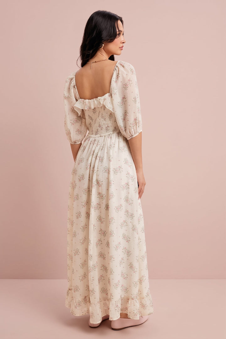 A woman wearing a cream-colored maxi dress with a delicate floral pattern, photographed from the back against a solid dusty pink studio backdrop.