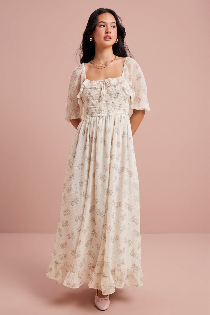 A woman modeling a cream-colored maxi dress with a delicate floral pattern, featuring a smocked bodice, ruffled short sleeves, and a square neckline, set against a solid dusty pink studio background.