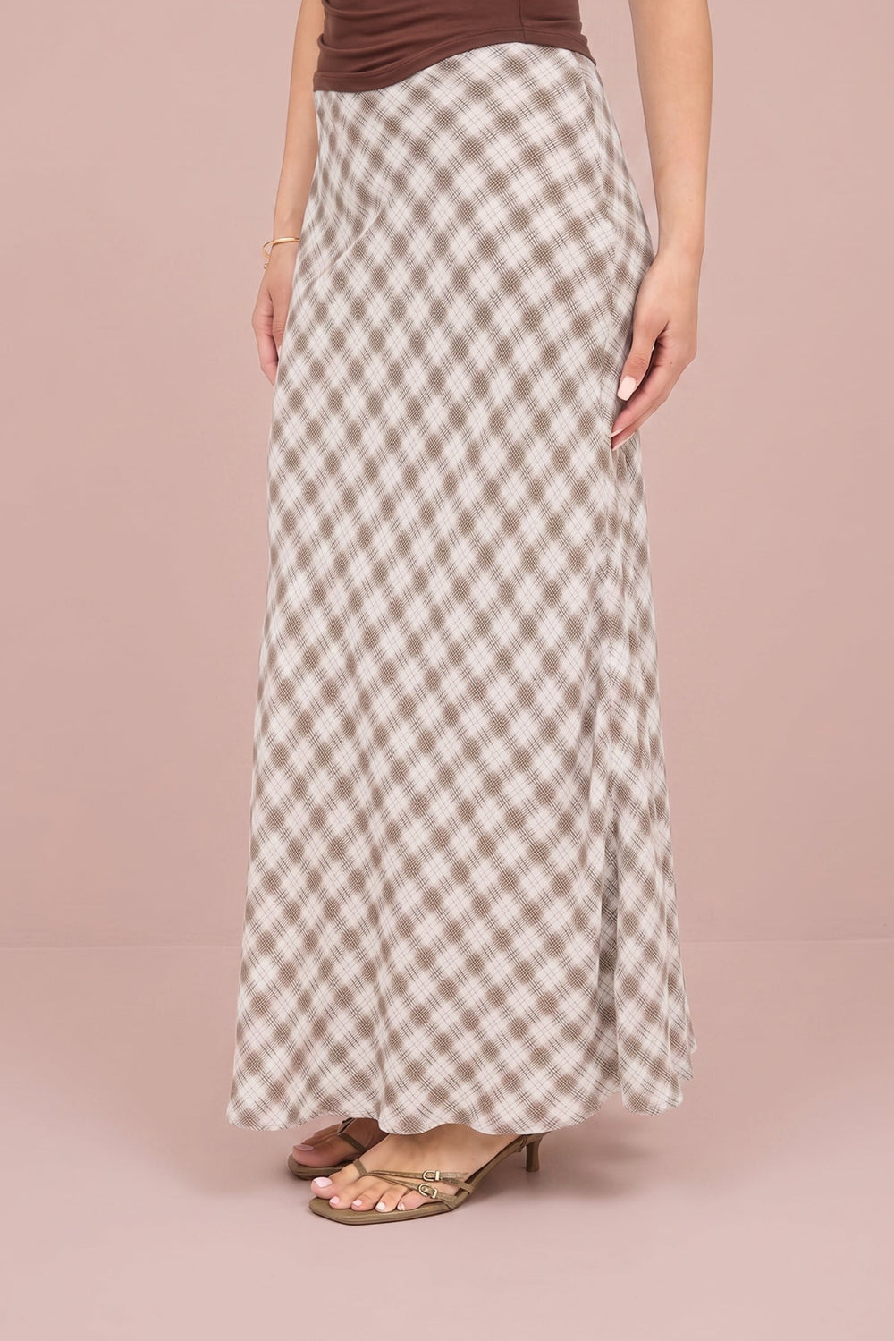 A woman wearing a brown and beige gingham check maxi skirt with a brown ruched top, standing against a solid dusty pink studio background.