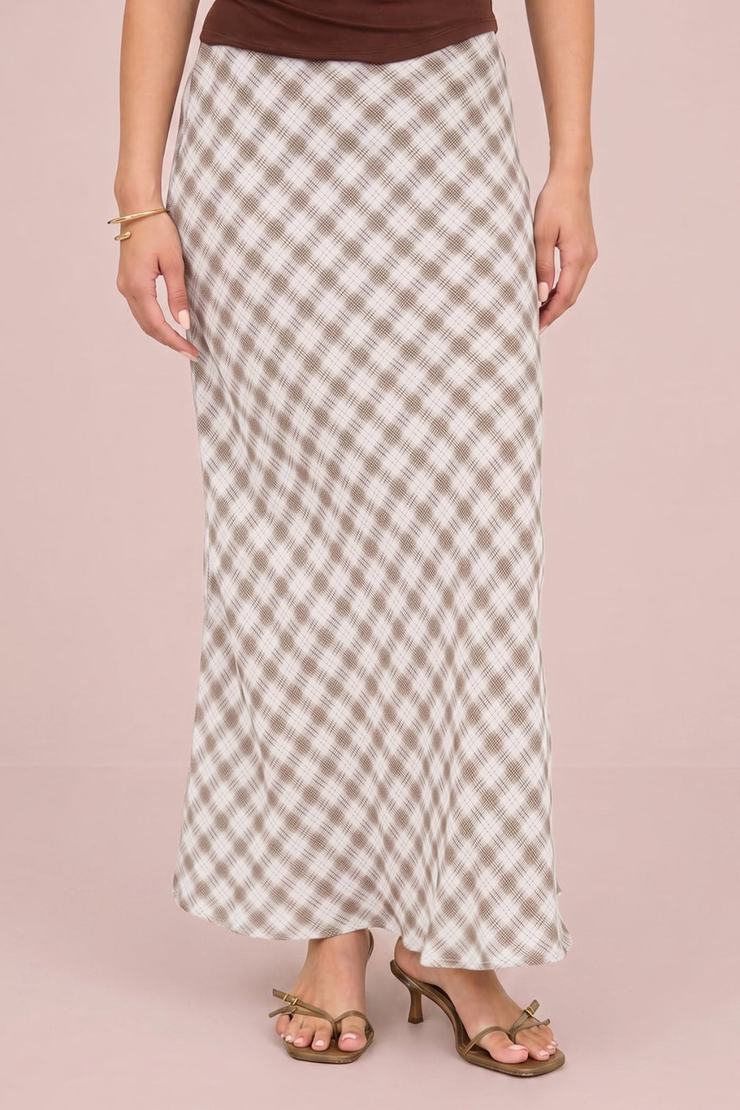 A woman models a long maxi skirt with a beige and white gingham check pattern, paired with a brown top and brown heeled sandals, set against a solid dusty pink background.