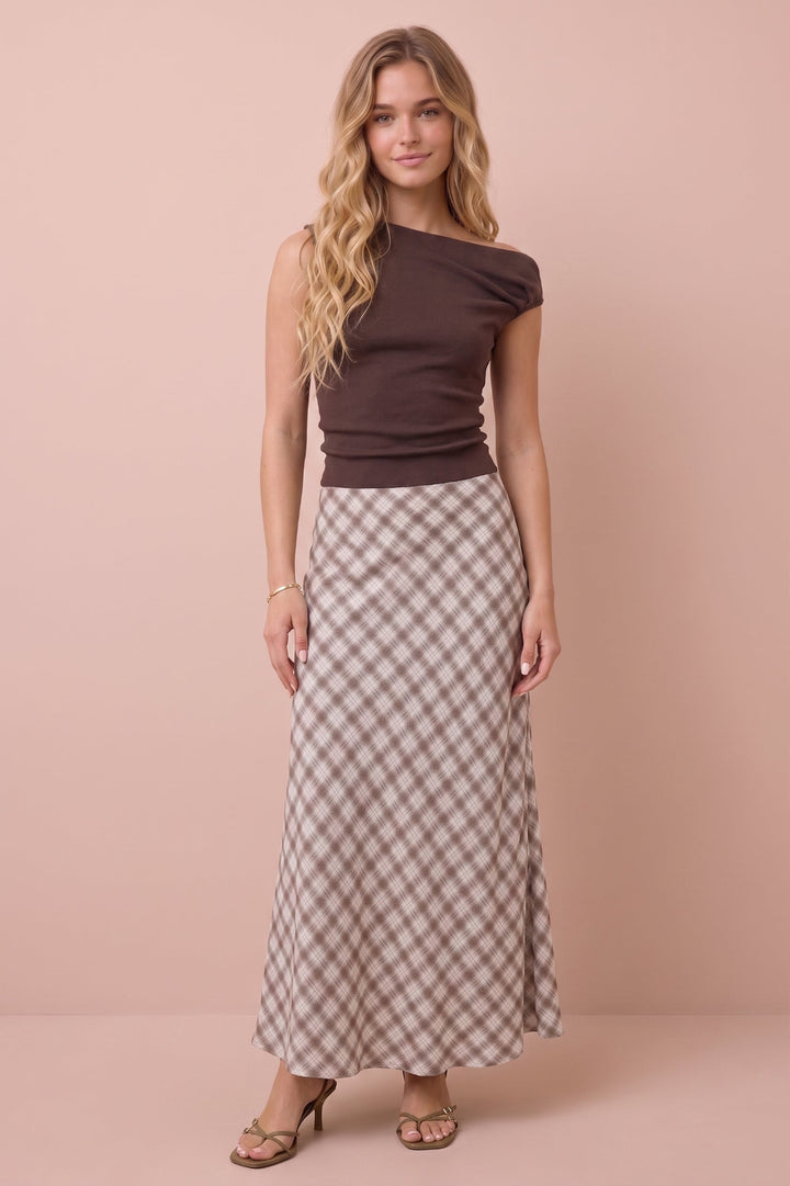A woman modeling a dark brown off-the-shoulder fitted top and a beige and brown gingham check maxi skirt, standing against a solid dusty pink studio background.