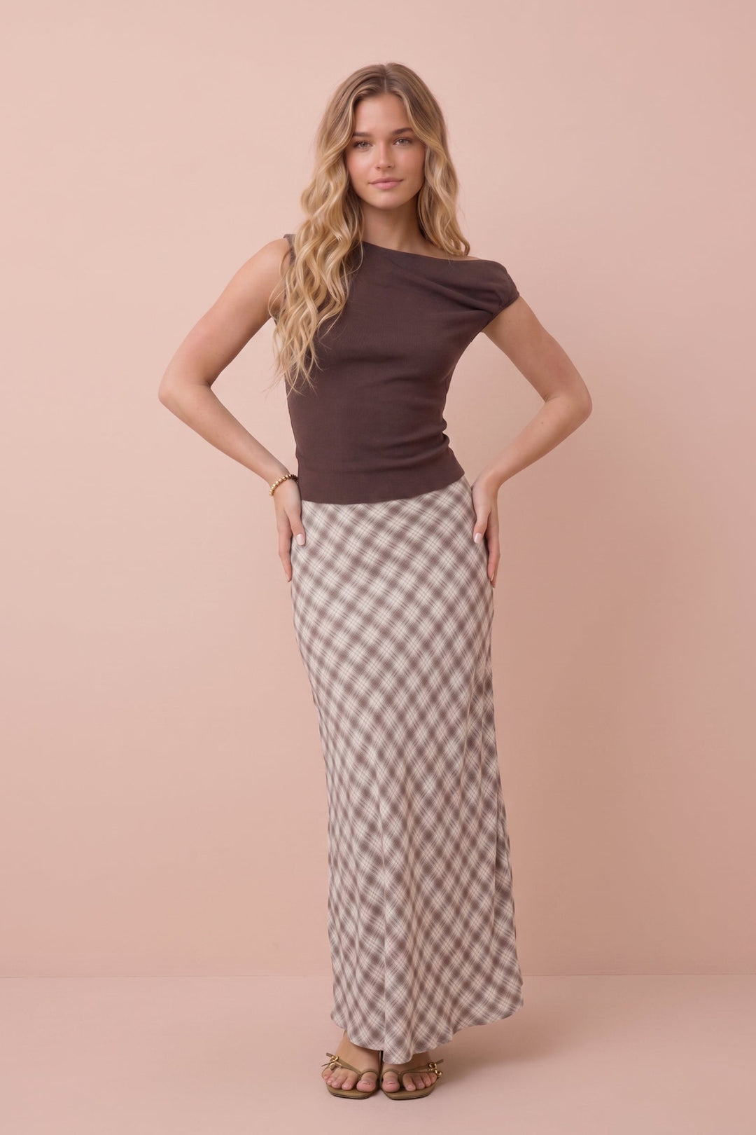 A woman modeling a dark brown off-the-shoulder top and a beige and brown checkered maxi skirt, standing against a solid pale pink studio backdrop.