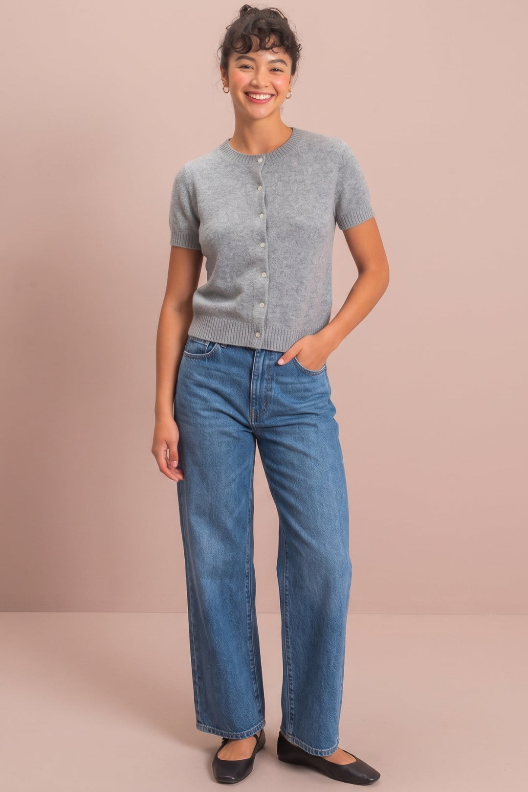 A woman stands against a solid dusty pink studio background wearing a light gray short-sleeve knit cardigan, blue wide-leg jeans, and black flats.