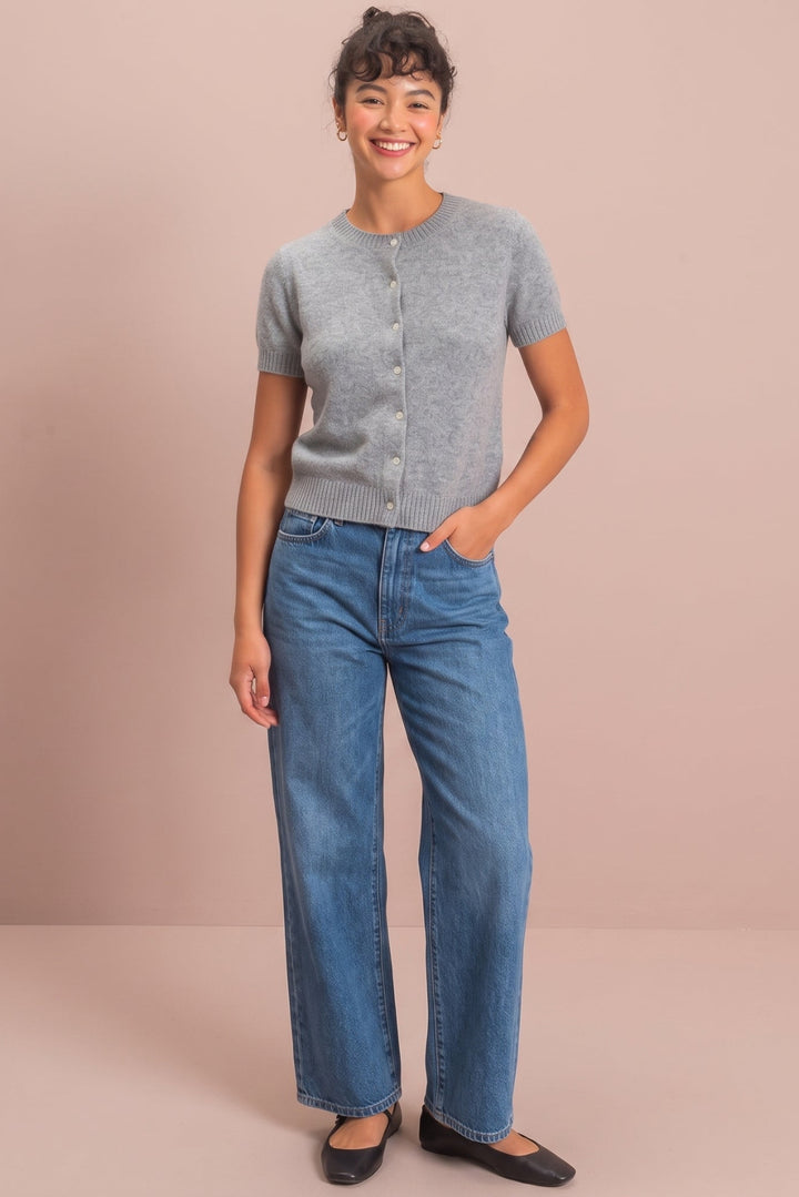 A woman stands against a solid dusty pink studio background wearing a light gray short-sleeve knit cardigan, blue wide-leg jeans, and black flats.