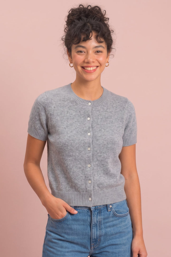 A smiling woman with curly dark hair stands against a solid dusty pink background, wearing a light gray short-sleeve button-up knit cardigan and blue jeans.