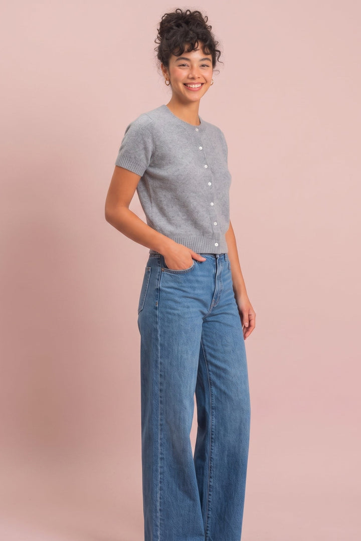 A woman modeling a light gray short-sleeve button-front cardigan and blue wide-leg jeans against a solid dusty pink studio background.