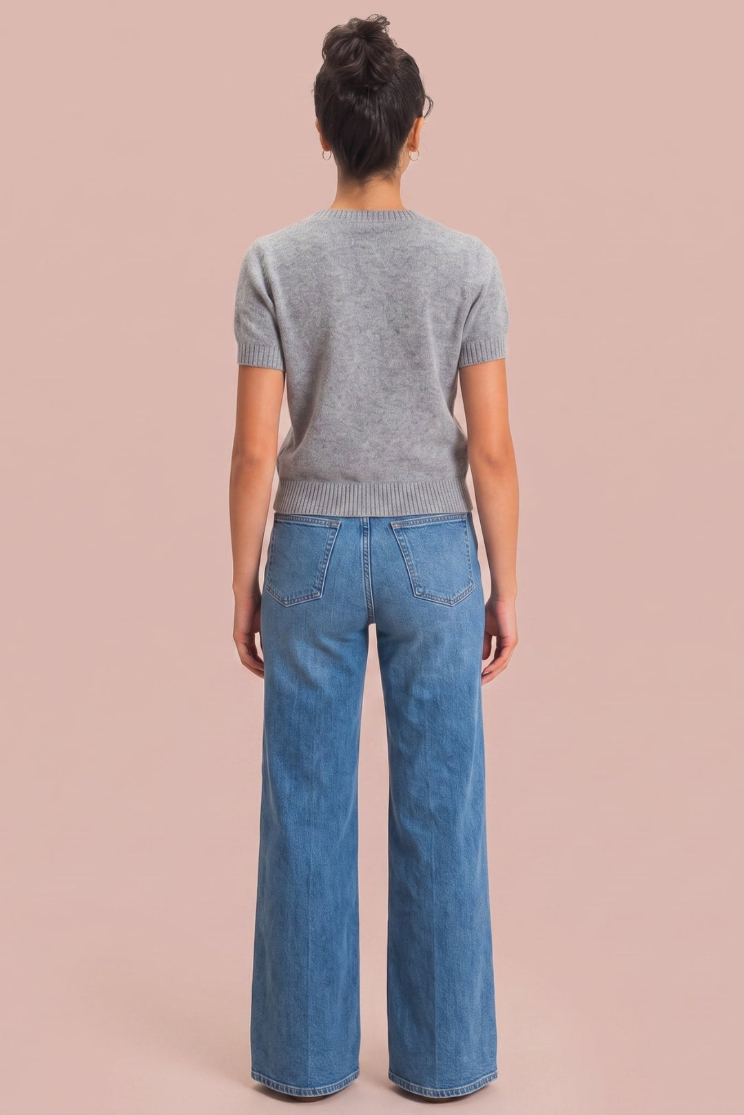 A model viewed from behind wearing a gray short-sleeve knit top and blue wide-leg jeans, standing against a solid dusty pink background.