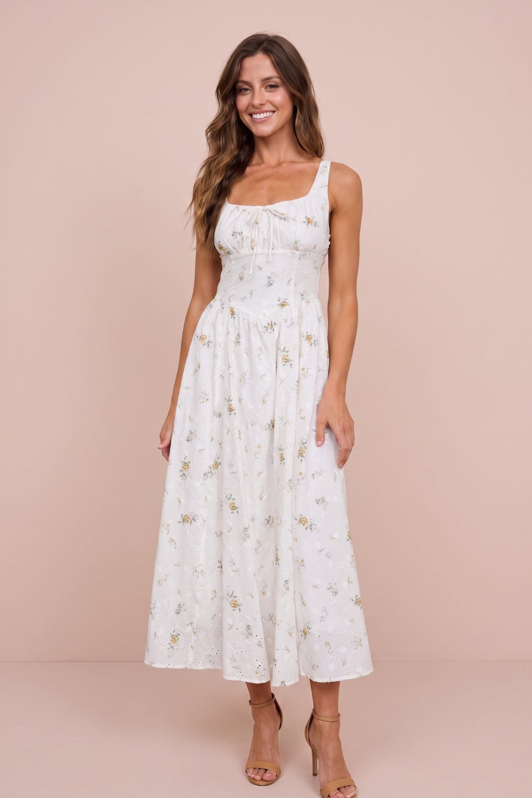 A woman modeling a white sleeveless midi dress with a delicate gold floral print, set against a solid pale pink studio backdrop.
