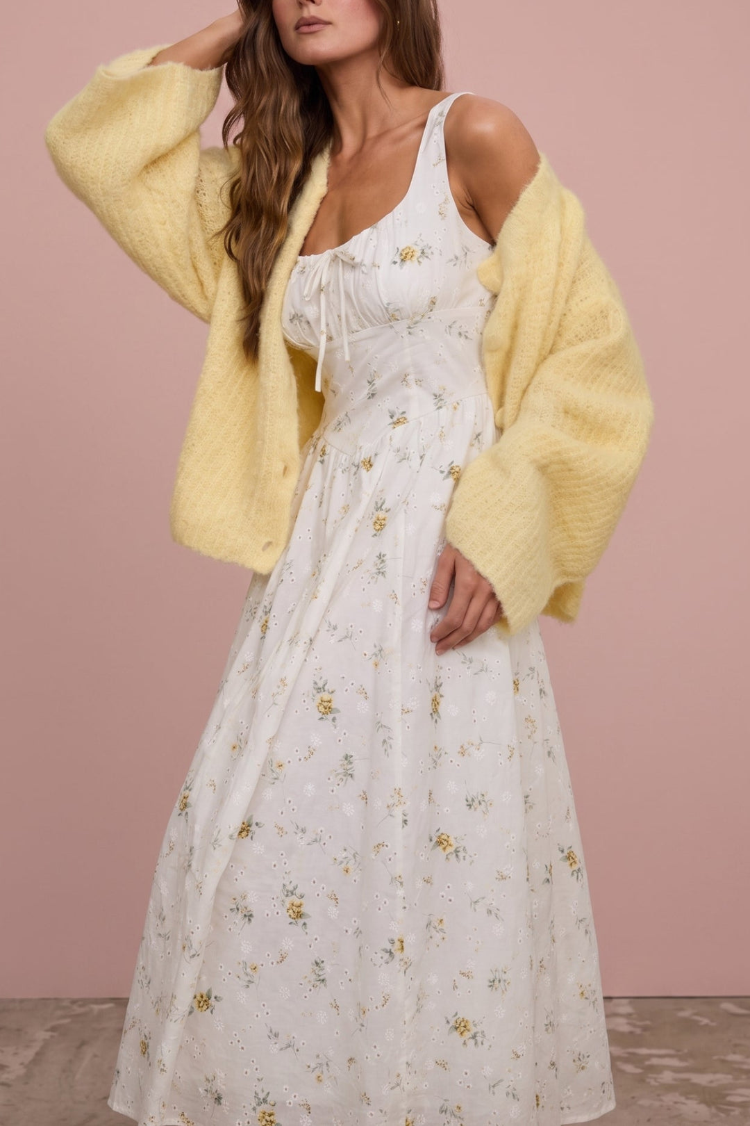 A woman stands against a dusty pink studio backdrop wearing a white floral midi dress with a yellow knit cardigan draped off her shoulders.