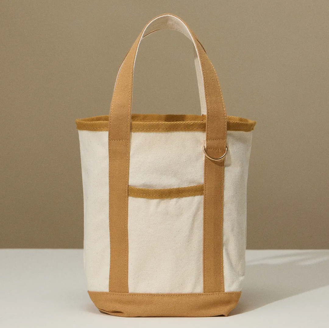 A small, rectangular canvas tote bag in a natural beige color with tan handles and trim, featuring a simple, minimalist design.