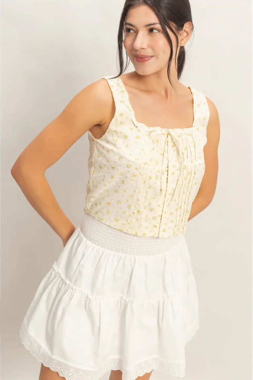 A young woman wearing a sleeveless white crop top with a floral print, paired with a white, ruffled skirt.