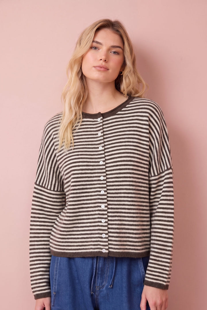 A woman modeling a brown and beige striped button-up cardigan sweater with dark brown trim, paired with blue jeans, set against a solid dusty pink background.