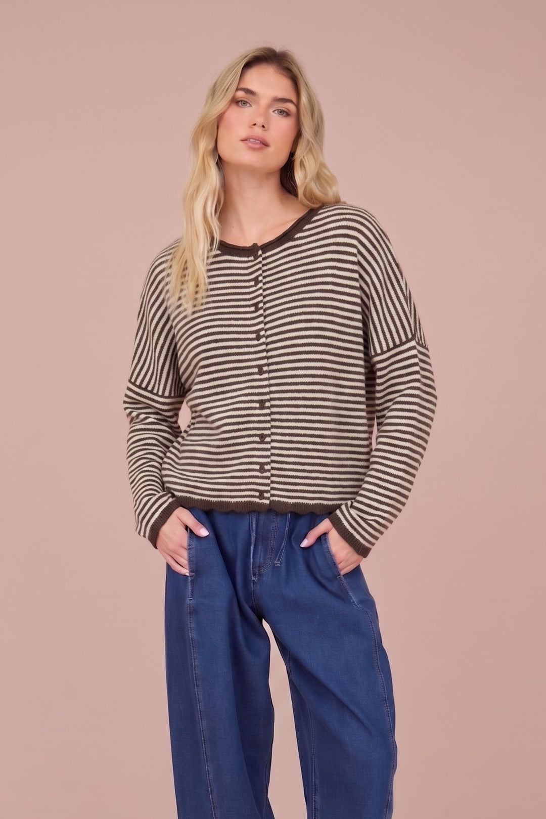 A woman modeling a black and beige striped knit long-sleeve top with button details, paired with wide-leg blue jeans and beige platform boots, set against a solid dusty pink studio background.