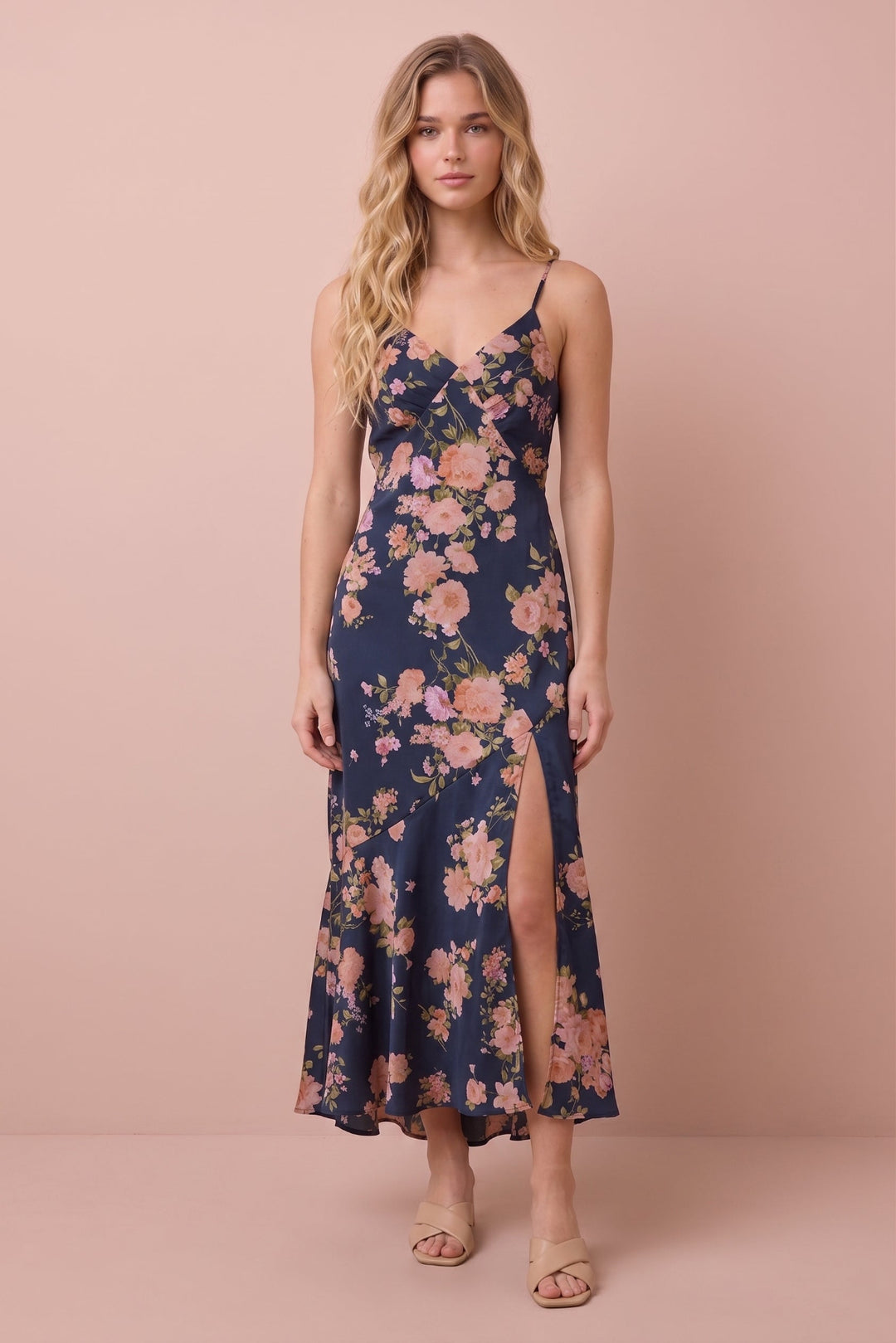 A woman modeling a navy blue maxi dress with a pink and peach floral print, spaghetti straps, and a thigh-high slit, set against a solid dusty pink studio background.
