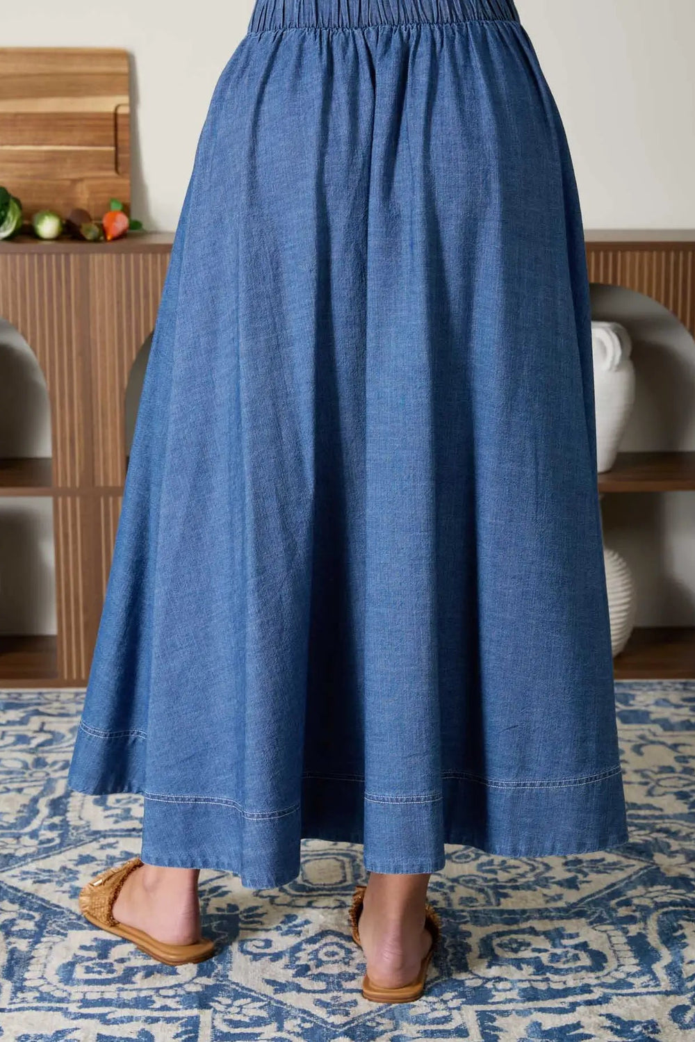 A mid-calf length denim skirt with a pleated design and elastic waistband, shown on a wooden floor with a decorative rug.