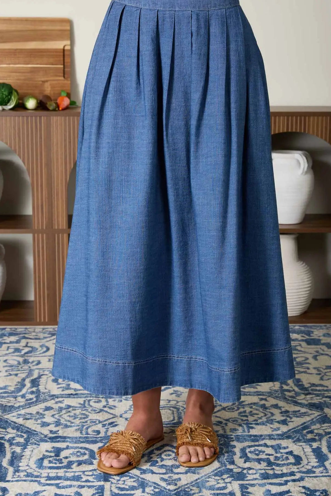 Navy blue pleated denim midi skirt with a flowing, high-waisted silhouette and stitched details, paired with woven slide sandals.