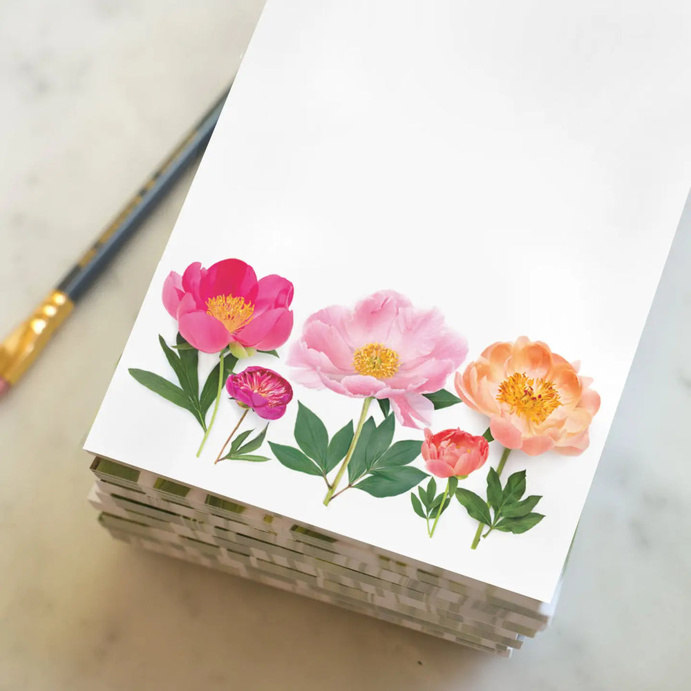 Vibrant peonies, pinks and oranges, against a white background on stacked notepads with a gold-toned pen.