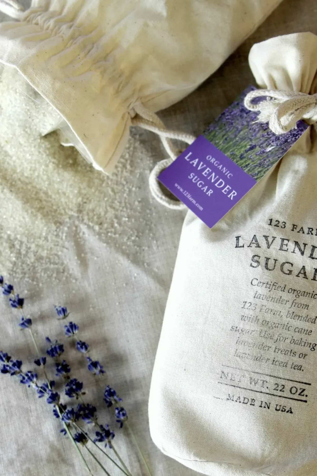 Close-up of a white fabric bag containing organic lavender-infused sugar, with a purple product tag and dried lavender sprigs.