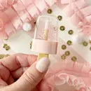 Popsicle-shaped lip gloss in pink and white with metallic gold flecks, displayed on a pink background with metallic confetti.