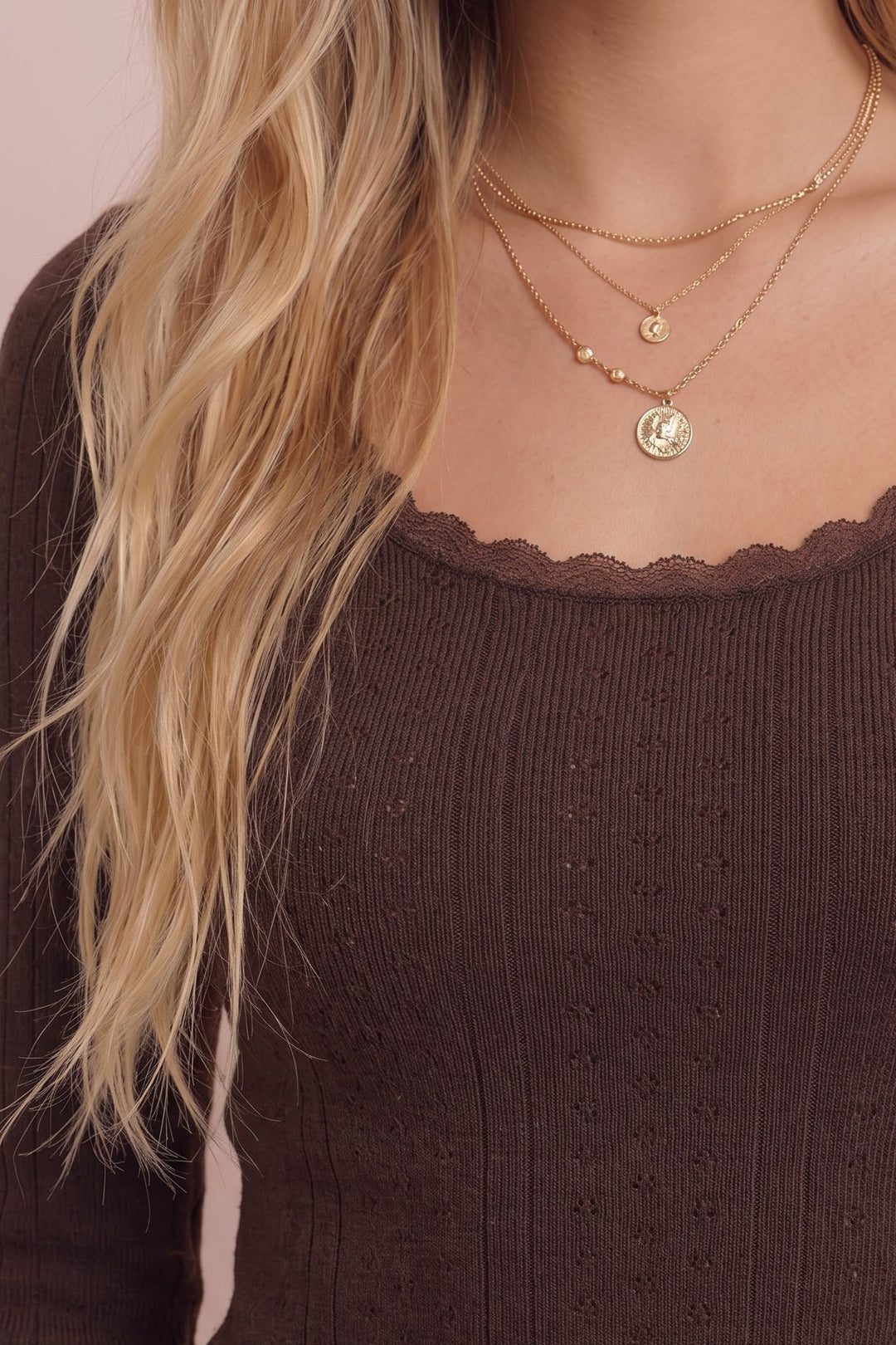 A close-up of a woman wearing a brown ribbed knit long-sleeve top with a scalloped scoop neckline, layered with delicate gold necklaces against a soft pink background.