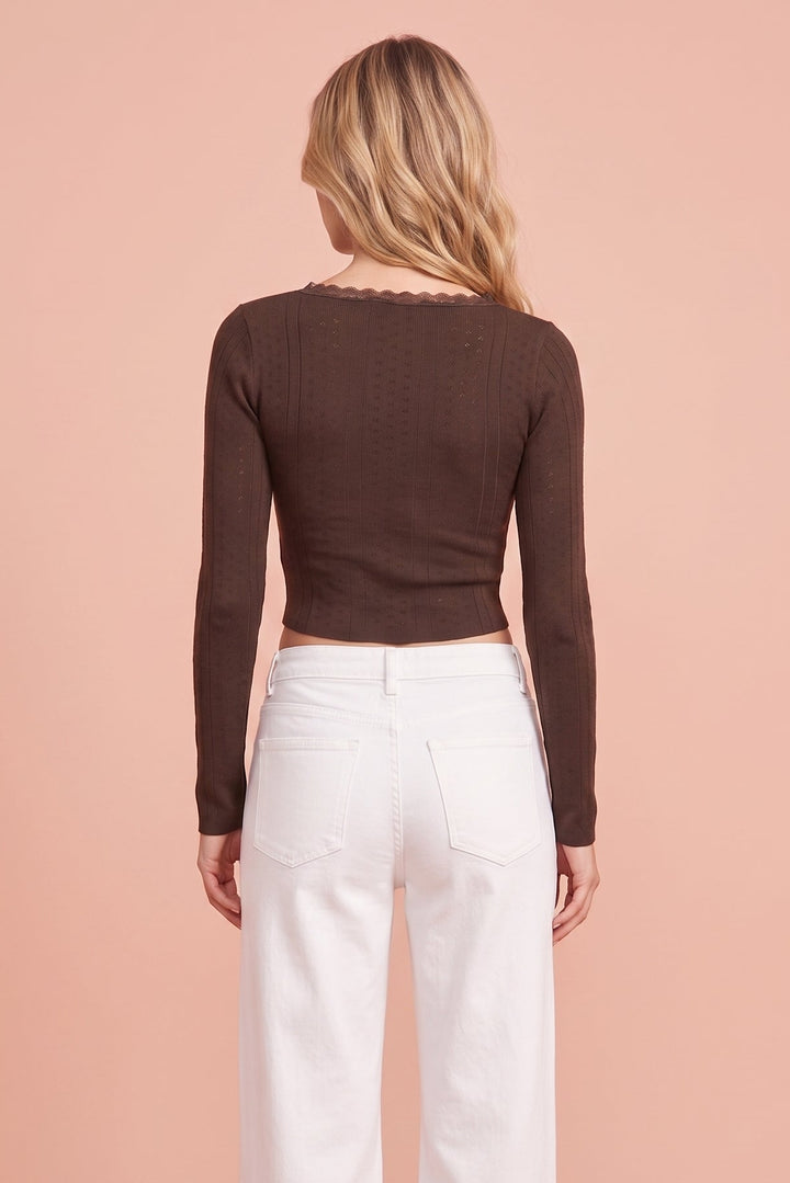A woman viewed from the back wearing a dark brown long-sleeve ribbed knit top with a scalloped neckline and white wide-leg pants, set against a solid peach-pink studio background.