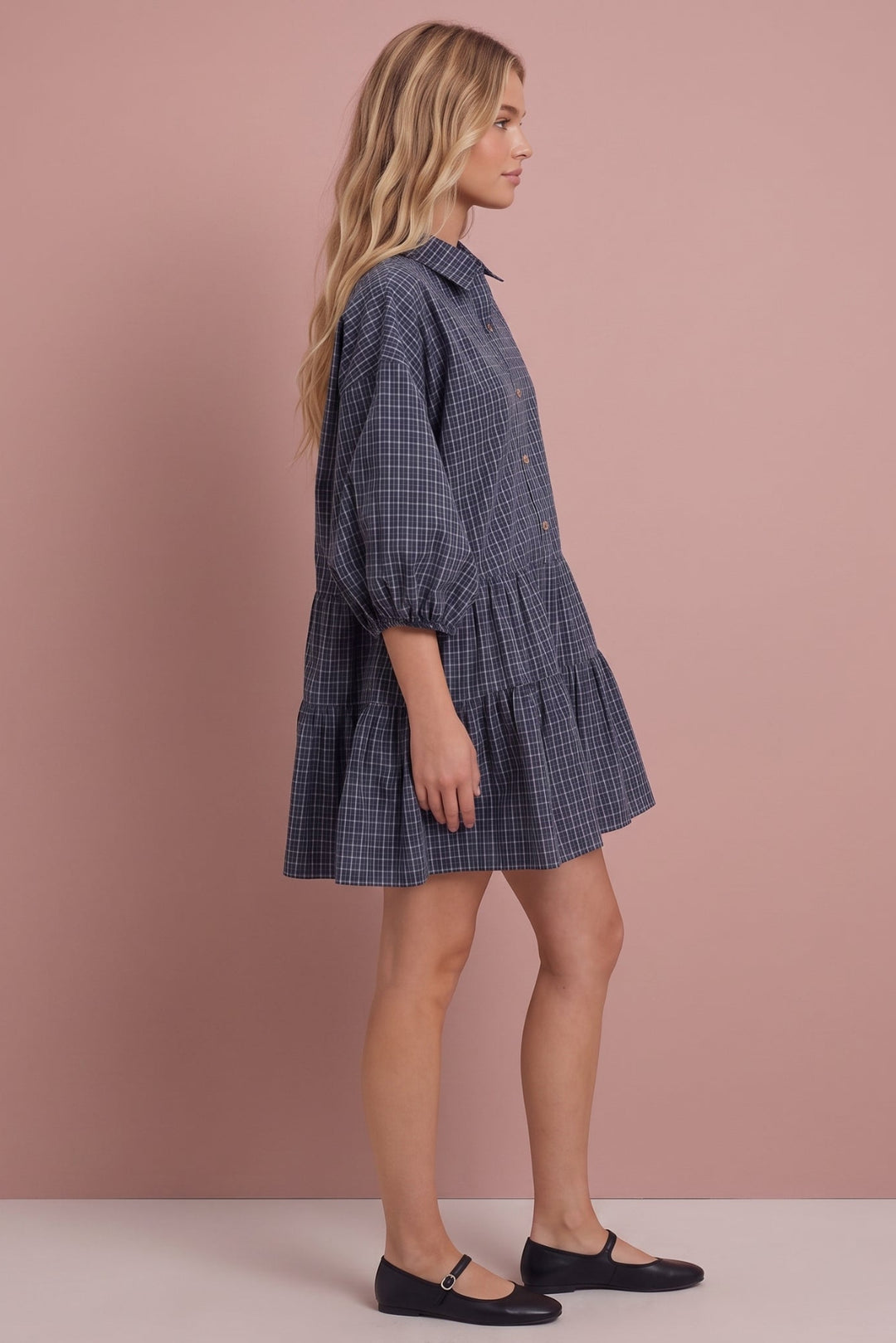 A woman modeling a navy and gray checkered mini shirt dress with puff sleeves, shown in profile against a solid dusty pink studio background.